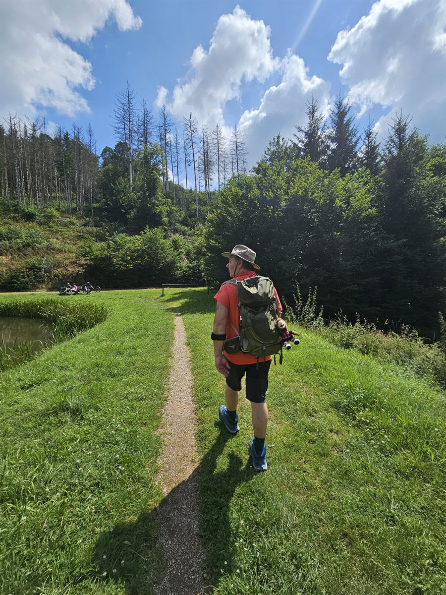 Georgios Katsiakoudis von hinten auf einem schmalen Pfad durch eine Wiese, mit Wanderhut und Rucksack, im Hintergrund Nadelwald.